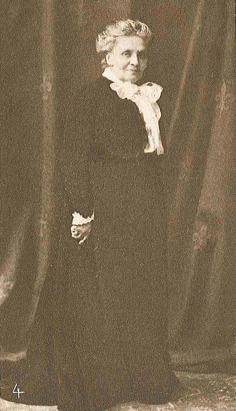 An elderly woman is standing in front of a curtain, wearing a dark dress with a white ruffled collar.
