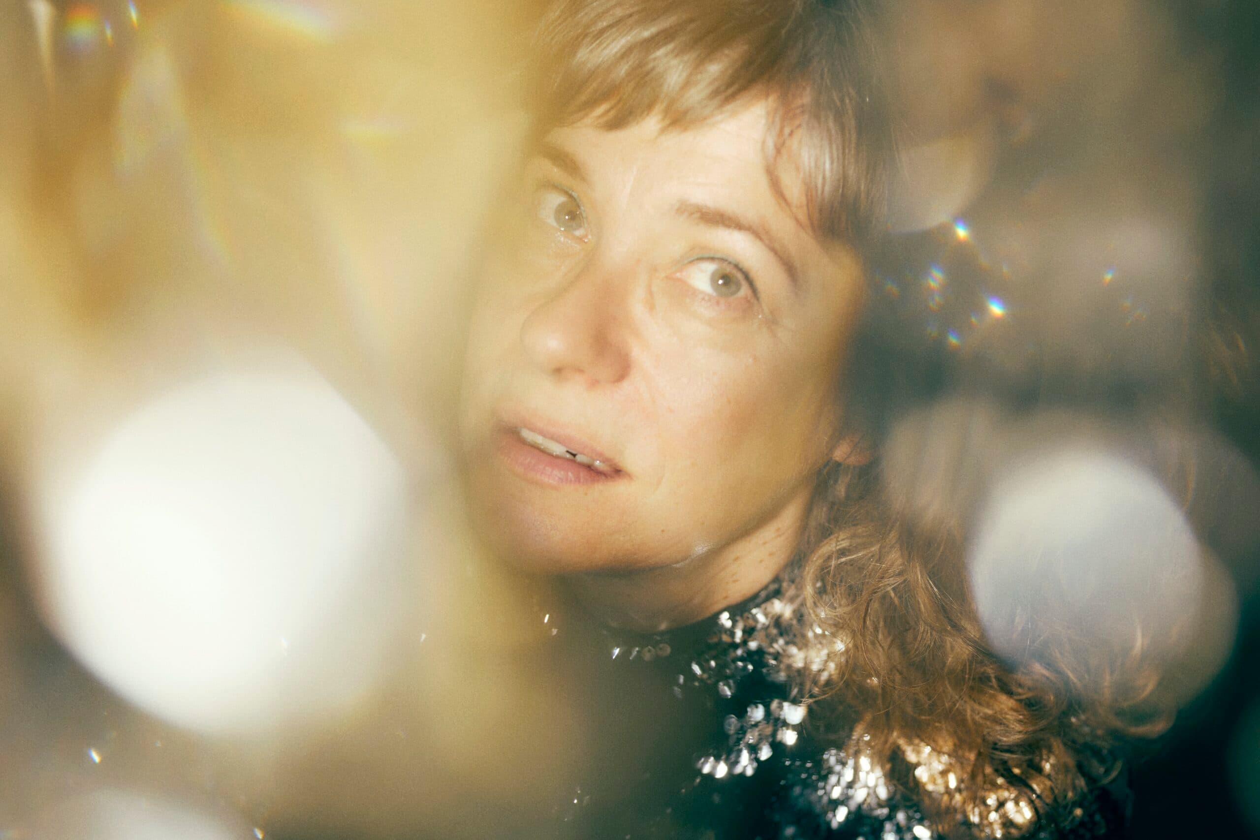 A woman with light brown hair looks upward, surrounded by soft, blurred light and reflections.