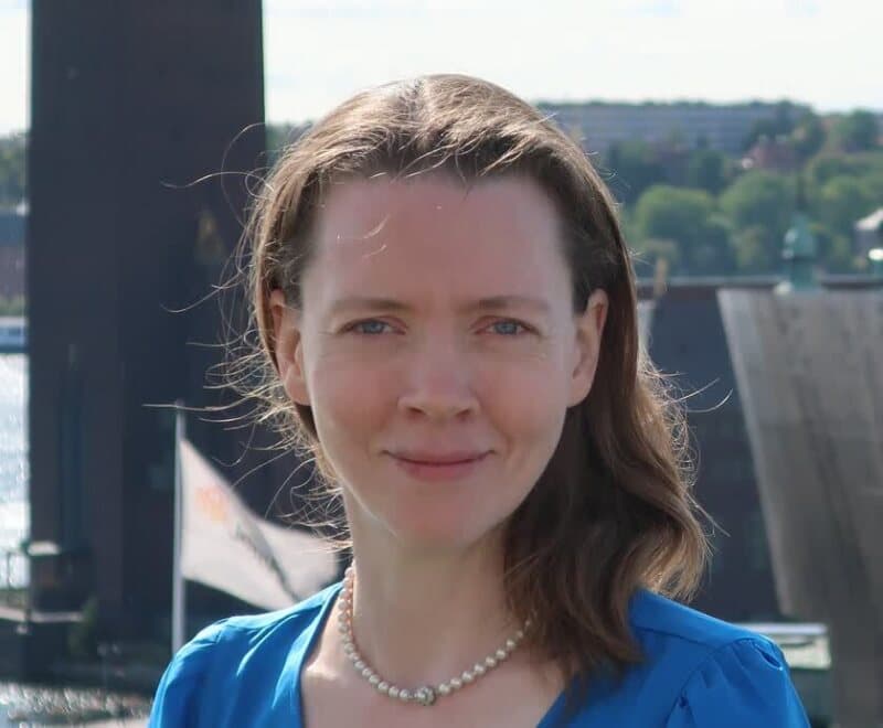 A person with shoulder-length hair and a pearl necklace is wearing a blue top, standing outdoors with buildings in the background.
