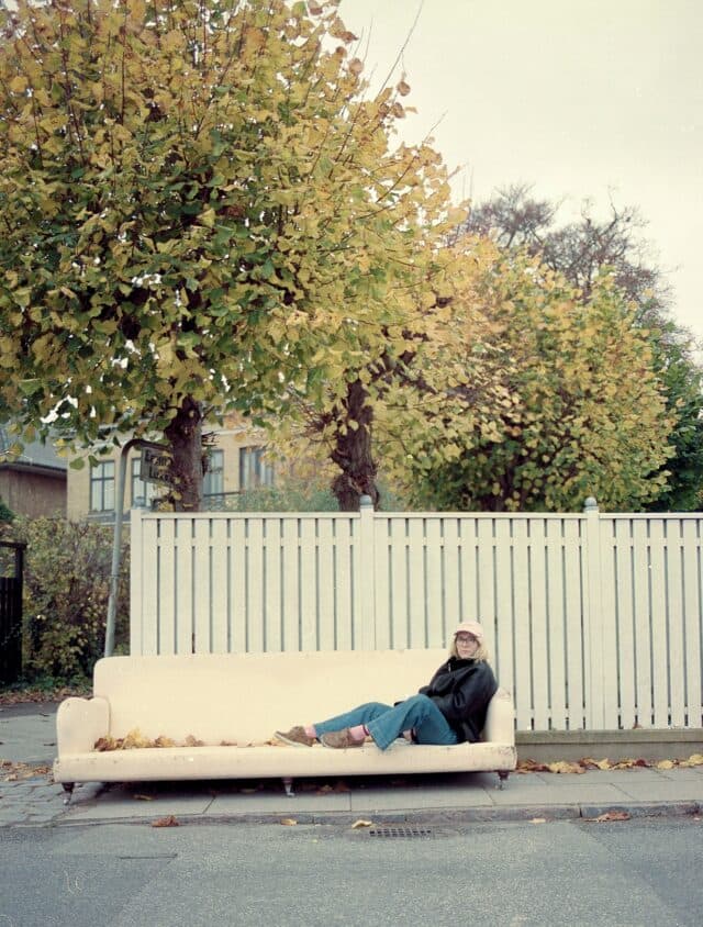 A person wearing a hat and glasses is sitting on a couch placed on a sidewalk, with autumn trees and a white fence in the background.