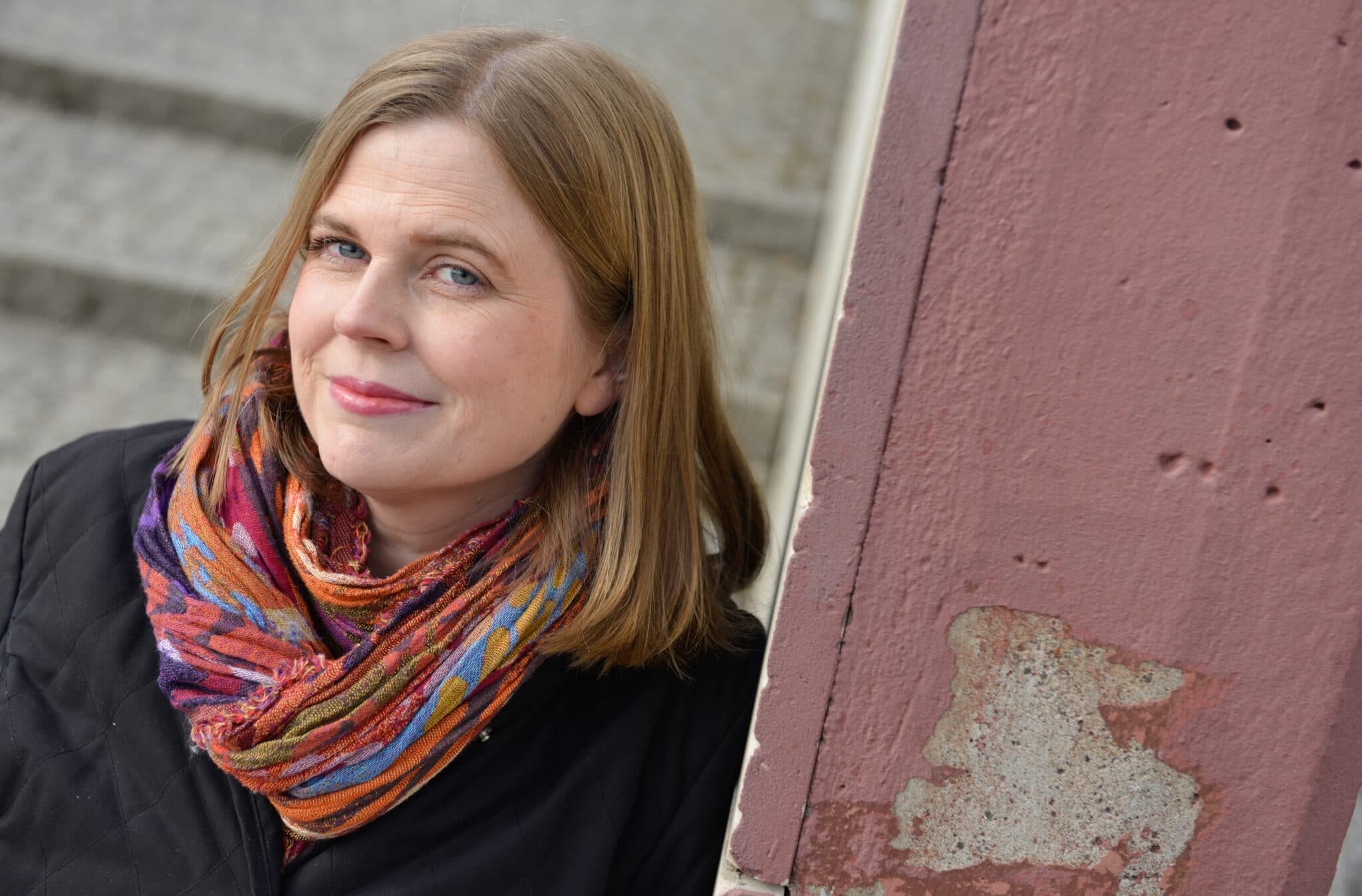 A woman with shoulder-length hair, wearing a colorful scarf and black coat, stands beside a weathered pink wall, smiling slightly.