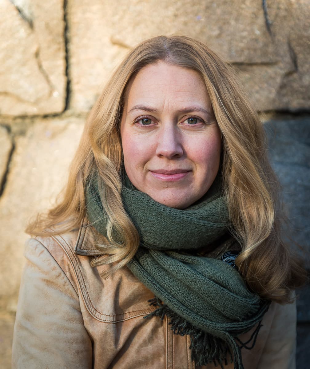 A woman with long blond hair wearing a green scarf and brown jacket stands in front of a stone wall.